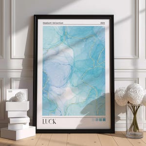 Op de afbeelding: Een ingelijste kunstdruk getiteld LUCK uit de Gradient Collection 2023. Het kunstwerk toont een abstract alcoholinktontwerp in tinten lichtblauw en turquoise, geaccentueerd met dunne, glinsterende gouden lijnen door de hele compositie heen.