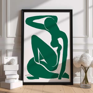 May include: A framed art print with a green silhouette of a seated figure on a white background. The artwork is in a black frame and is signed in the lower right corner. The print is displayed on a wooden floor next to a vase of white flowers.