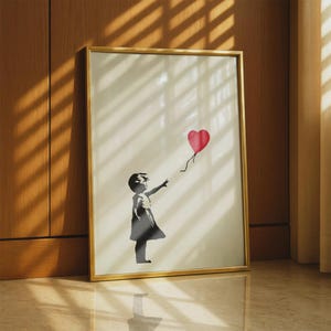 Könnte beinhalten: Ein gerahmter Kunstdruck mit der schwarzen Silhouette eines Kindes, das nach einem leuchtend roten, herzförmigen Luftballon greift. Das Motiv befindet sich auf einem cremeweißen Hintergrund und ist in einem dünnen, goldfarbenen Rahmen gefasst.