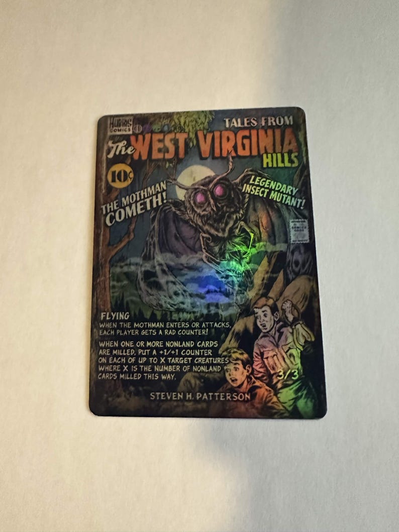 May include: A collectible card featuring a comic book style illustration. The card reads "Tales From The West Virginia Hills" with a Mothman creature and the text "The Mothman Cometh!" and "Legendary Insect Mutant!"