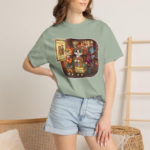 以下が含まれることがあります： セージグリーンの半袖Tシャツ。ショップにいる3匹の擬人化された猫のグラフィックイラストが描かれています。猫たちは「Human Doll Class Room 2B」と書かれた看板と、「Woodland Crafts」とラベル付けされたトートバッグと一緒に描かれています。