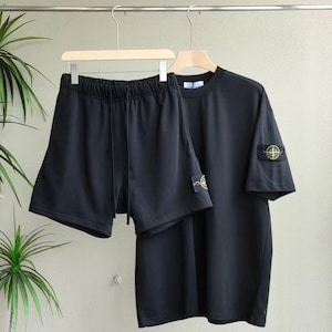 以下が含まれることがあります： 黒色のショートパンツとTシャツのセット。ショートパンツはドローストリングウエストです。Tシャツは半袖で、袖に小さなパッチが付いています。どちらのアイテムも木製のハンガーに吊るされています。