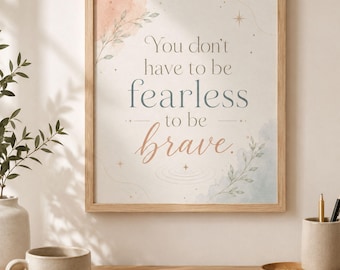 May include: A framed wall art print featuring the text 'You don't have to be fearless to be brave.' The design includes soft watercolor accents in peach and blue, delicate leaf illustrations, and small star symbols on a light background.
