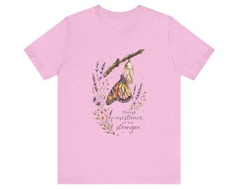 May include: Light pink t-shirt featuring a watercolor illustration of a monarch butterfly emerging from a chrysalis on a branch, surrounded by flowers and foliage. The text "Through resistance, we rise stronger" is below the butterfly.