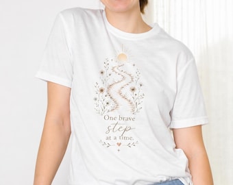 May include: White t-shirt and baseball cap with the text "One brave step at a time." The shirt features a winding path design with floral accents and a sun graphic. The cap has the same text.