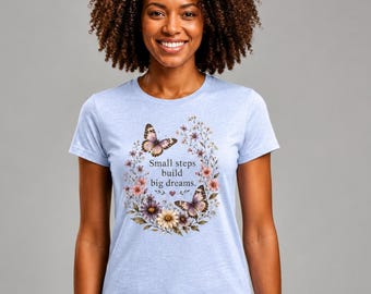 May include: A light blue t-shirt featuring a floral and butterfly design. The design includes the text "Small steps build big dreams." The butterflies are brown and white, and the flowers are various colors. The shirt is a crew neck.