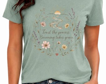 May include: Sage green t-shirt with a floral wreath design. The wreath surrounds a sun and river graphic, with the text "Trust the process. Becoming takes grace." The shirt is paired with blue jeans.