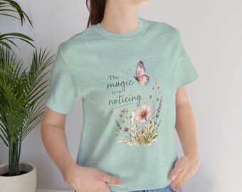 May include: A light green t-shirt featuring a floral and butterfly design. The design includes a pink flower, purple wildflowers, and a butterfly. The text "The magic is in noticing" is printed above the floral design.
