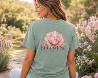 May include: A sage green short-sleeve t-shirt featuring a large pink lotus flower graphic on the back. Below the flower, the text reads BECOMING in a dark serif font, with the smaller text TRUST THE PROCESS printed underneath the word.