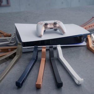 Puede incluir: Una consola de juegos blanca y negra con un mando blanco encima, rodeada de soportes de consola de varios colores. Los soportes son de color plata, cobre, negro y otros colores metálicos. La consola y los accesorios se muestran sobre una superficie gris.