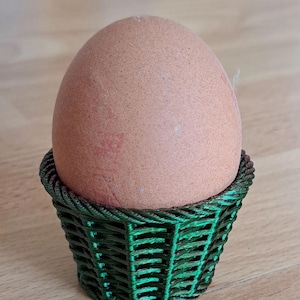Puede incluir: Un huevo marrón en un huevera verde tejida. El huevo tiene un aspecto moteado y una marca rojiza. La huevera está hecha de un material verde oscuro y tiene un diseño de tejido de cesta. La huevera mide aproximadamente 6 cm de alto.