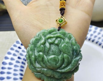 May include: A circular green jade pendant carved into a detailed floral shape with layered petals and a textured center. The pendant is attached to a dark brown cord featuring a gold-toned metal accent piece with a small green stone inlay.
