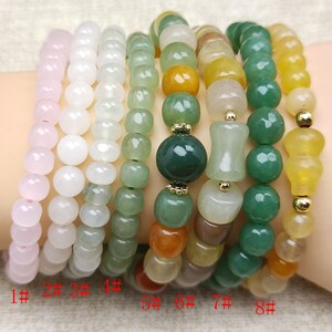 May include: Eight beaded bracelets in various colors, including pink, white, green, yellow, and brown. The bracelets feature different bead shapes and sizes, with some having gold-colored accents. The bracelets are displayed on a light-colored surface.