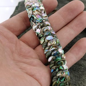 May include: A strand of iridescent abalone shell beads in a variety of green, blue, and brown tones. The beads are round and have a smooth, polished finish.