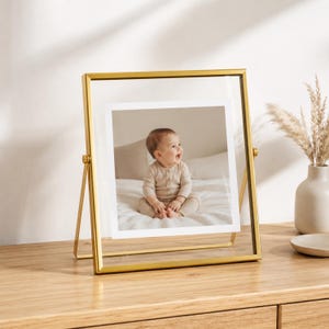 May include: A gold-toned metal floating picture frame with a square glass pane holding a photograph of a baby wearing a beige long-sleeved onesie. The frame features a hinged stand design, allowing it to tilt, and is displayed on a wooden surface.