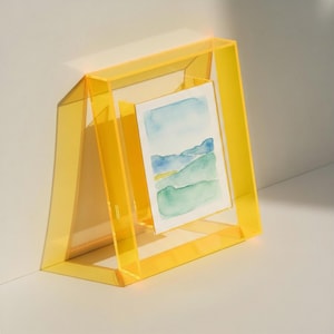 May include: A translucent yellow acrylic picture frame with a trapezoidal base, holding a small rectangular watercolor painting that depicts a landscape with blue mountains and green rolling hills under a light blue sky.