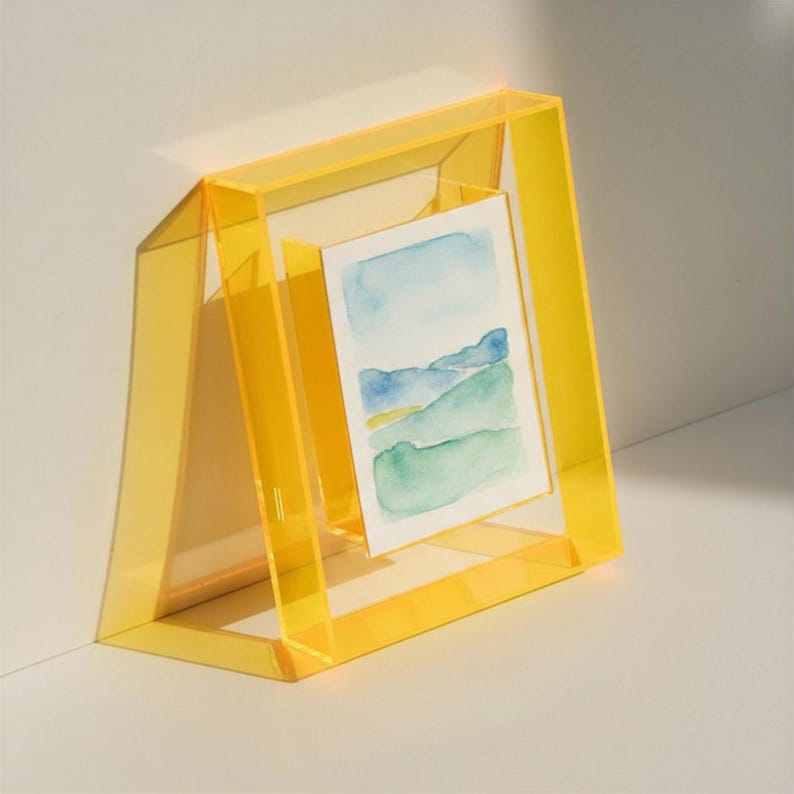 May include: A translucent yellow acrylic picture frame with a trapezoidal base, holding a small rectangular watercolor painting that depicts a landscape with blue mountains and green rolling hills under a light blue sky.