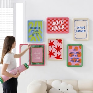 May include: A collection of framed art prints with colorful designs and text. The prints include phrases like "Radiate Positivity," "Wild Child," and "Crying Corner." The frames have various colors, including pink, green, and blue.