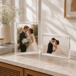 May include: Two clear acrylic block photo frames displaying wedding photographs. The left frame is vertical, showing a couple standing together, while the right frame is horizontal, showing the same couple embracing. The frames have silver hardware.