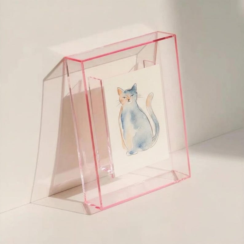 May include: A clear acrylic picture frame with a pink border containing a white card featuring a watercolor illustration of a seated cat with blue and grey fur tones. The frame is angled and stands upright, showcasing the artwork clearly.