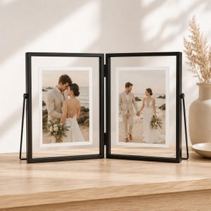 Puede incluir: Un marco de fotos doble articulado de metal negro con dos paneles de vidrio transparente. Cada panel contiene una fotografía vertical de una pareja en una playa. El marco presenta bordes negros finos y un diseño plegable para mantenerse en pie.