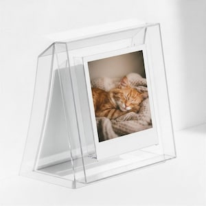 May include: A clear acrylic easel-style photo frame holding a vertical photograph of an orange tabby cat sleeping on a textured beige blanket. The frame features a slanted, transparent design that supports the photo in an upright position.