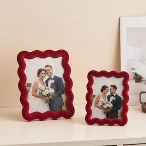 Puede incluir: Dos marcos de fotos de terciopelo rojo con borde ondulado, cada uno con una foto de boda. Los marcos son de diferentes tamaños y están sobre una superficie blanca. Las fotos muestran a los novios.