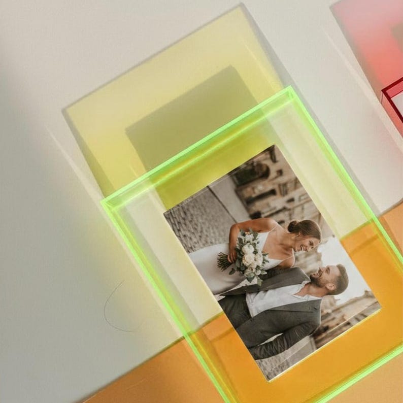 May include: A rectangular neon yellow acrylic picture frame holding a photograph of a couple. The photograph shows a person in a white dress holding a bouquet and a person in a grey suit standing together outdoors on a stone path.