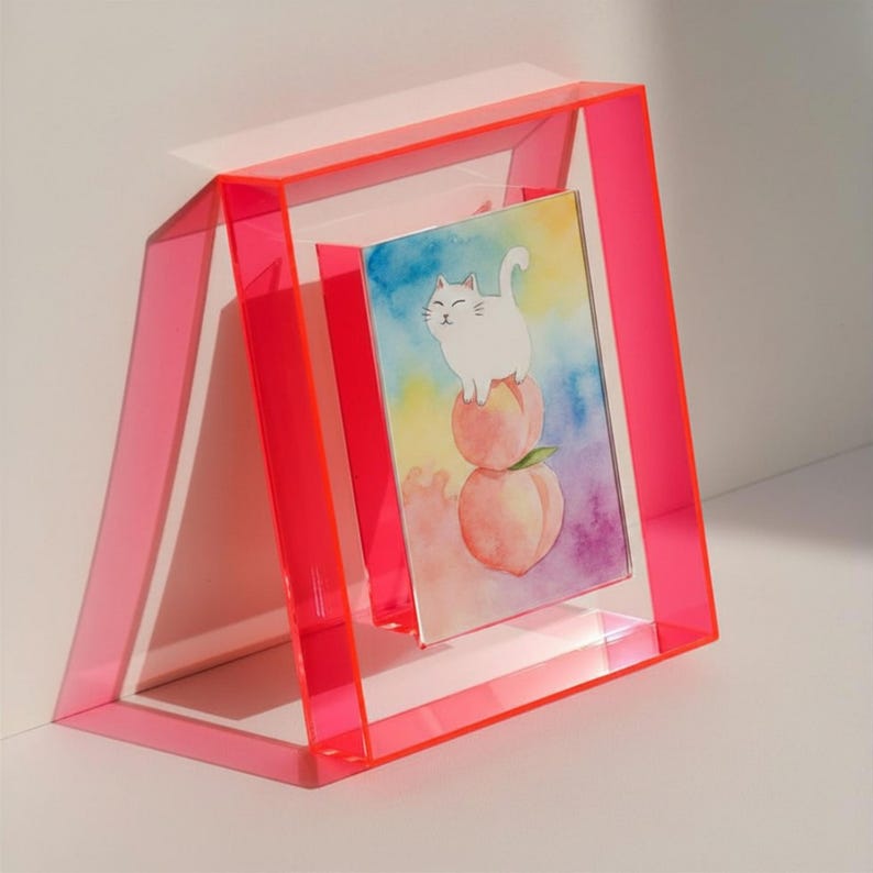May include: A rectangular, neon pink acrylic frame holds a vertical print featuring a white cat standing on top of two stacked peaches. The background of the print is a soft, watercolor-style gradient of blue, yellow, and purple hues.