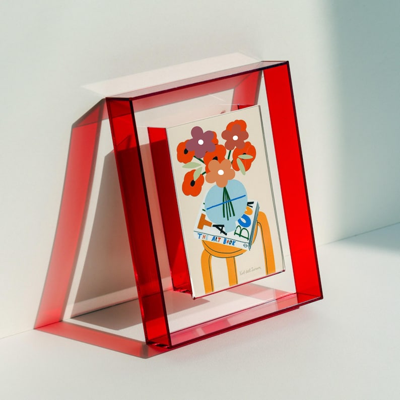 May include: A rectangular, transparent red acrylic picture frame holding a vertical print. The print features a colorful illustration of a bouquet of flowers in a light blue vase, resting on a book titled THE ART BOOK, all set on a yellow stool.