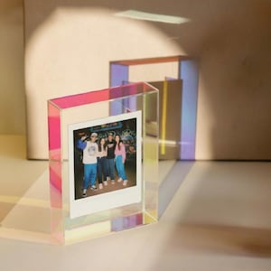 May include: A rectangular, iridescent acrylic photo frame with a clear center holding a square instant photograph of three people standing together. The frame reflects light in shades of pink, blue, and yellow, creating a colorful glow.