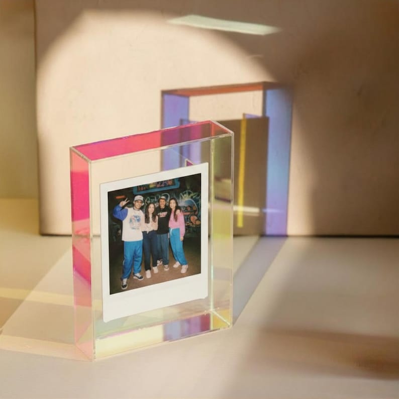 May include: A rectangular, iridescent acrylic photo frame with a clear center holding a square instant photograph of three people standing together. The frame reflects light in shades of pink, blue, and yellow, creating a colorful glow.