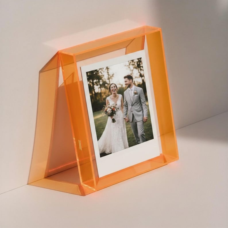 May include: A translucent neon orange acrylic easel-style picture frame holding a photograph of a person in a white wedding dress and a person in a grey suit standing together outdoors in a grassy area with trees in the background.