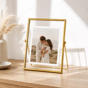 May include: A rectangular floating picture frame with a thin gold metal border and a pivoting stand. The frame holds a photograph of two adults and a child standing in a grassy field. The frame is constructed with clear glass panels.