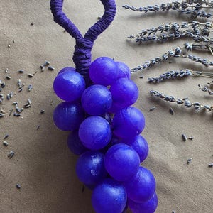 以下が含まれることがあります： 紫色のブドウの形をした手作りの装飾品。ブドウは濃い紫の色で、巻き紐で作られたループハンドルが付いています。乾燥したラベンダーの小枝と種が、茶色の表面のブドウの周りに散らばっています。