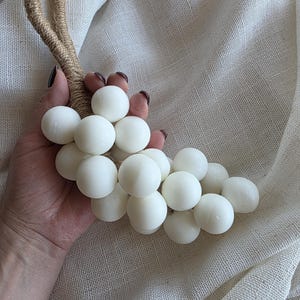 Może przedstawiać: Dekoracyjny kiść białych kul, przypominających winogrona, z pętlą z naturalnej liny. Kule są gładkie i równomiernie wielkości. Uchwyt z liny wykonany jest z ciasno zwiniętych, jasnobrązowych włókien. Tło to teksturowana, kremowa tkanina.