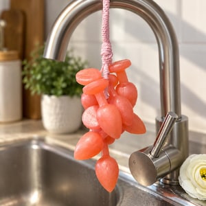 May include: A cluster of translucent, coral-pink soap pieces shaped like small teardrops or bulbs, strung together on a light pink braided cord. The soap cluster hangs from a curved, brushed stainless steel kitchen faucet fixture.