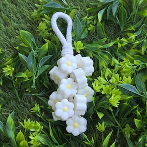 May include: A white macrame cord loop attached to a cluster of small, white, flower-shaped beads with yellow centres. The beads are arranged in a dense, hanging formation, creating a floral-inspired accessory with a simple, clean design.