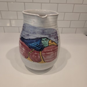 May include: A white ceramic pitcher with a hand-painted illustration of a bird. The bird has a dark blue body, a light green head, a yellow beak, and yellow legs. The background of the illustration features a purple sky and a red ground.