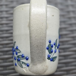 Op de afbeelding: Een gespikkelde gebroken witte keramische mok met een textuurrijk handvat en een handgeschilderd ontwerp van groene bladeren en verhoogde blauwe bessen. De mok heeft een cilindrische vorm met een makersmerk bij de basis van het handvat.