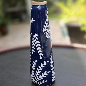 May include: A tall, slender ceramic vase with a glossy dark blue glaze and a hand-painted white fern leaf pattern. The vase features a narrow neck with a light beige, unglazed rim and a slightly tapered, cylindrical body that widens toward the base.