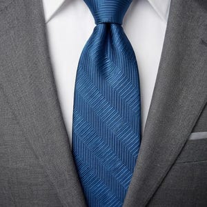 Peut inclure: Gros plan d'une cravate bleue à motif chevrons, nouée sur une chemise blanche et une veste de costume grise. La texture et la couleur de la cravate sont mises en valeur, suggérant une tenue formelle ou professionnelle.