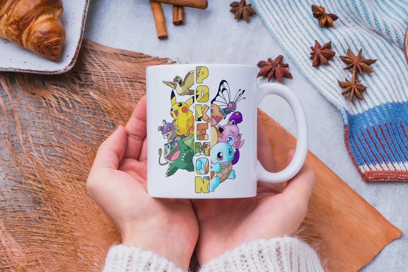 May include: White ceramic mug featuring a colorful design of various Pokemon characters and the word "Pokemon" in yellow and blue. The mug has a large handle and is held by a person wearing a white sweater. The mug is a perfect gift for fans of the popular franchise.
