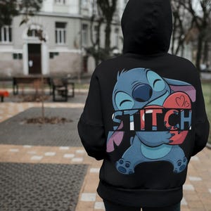 May include: Black hooded sweatshirt featuring a large graphic of the character Stitch from Lilo & Stitch. Stitch is blue with large ears, holding a red heart. The word "STITCH" is printed in white across the design.