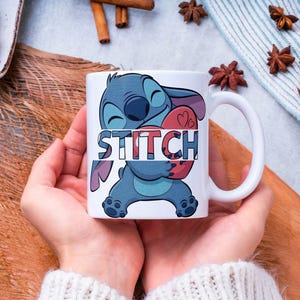 May include: White ceramic mug featuring the character Stitch from Lilo & Stitch, holding a red heart. The word "STITCH" is printed across the character. The mug has a white handle and is held by a person.