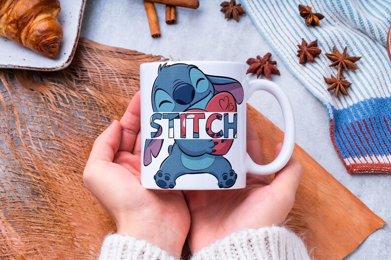 May include: White ceramic mug featuring the character Stitch from Lilo & Stitch, holding a red heart. The word "STITCH" is printed across the character. The mug has a white handle and is held by a person.