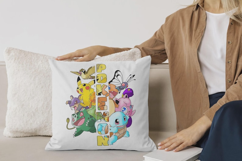 May include: White square pillow featuring a colorful graphic of various Pokemon characters, including Pikachu, Bulbasaur, and Squirtle. The word "Pokemon" is vertically printed in yellow and orange. The pillow is on a white couch.