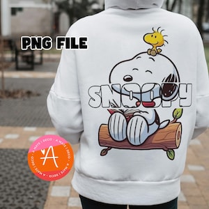 Puede incluir: Una sudadera con capucha blanca que presenta un gráfico en la parte posterior del personaje de dibujos animados Snoopy sentado en un tronco marrón mientras lee un libro, con el pájaro amarillo Woodstock posado sobre la cabeza de Snoopy.