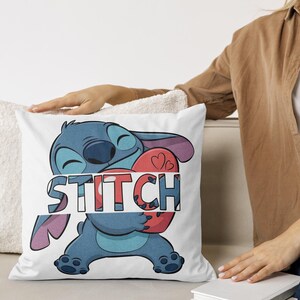 May include: White square pillow featuring a blue cartoon character holding a red heart with two small hearts. The word "STITCH" is printed across the character in a colorful, blocky font. The pillow is on a white couch.