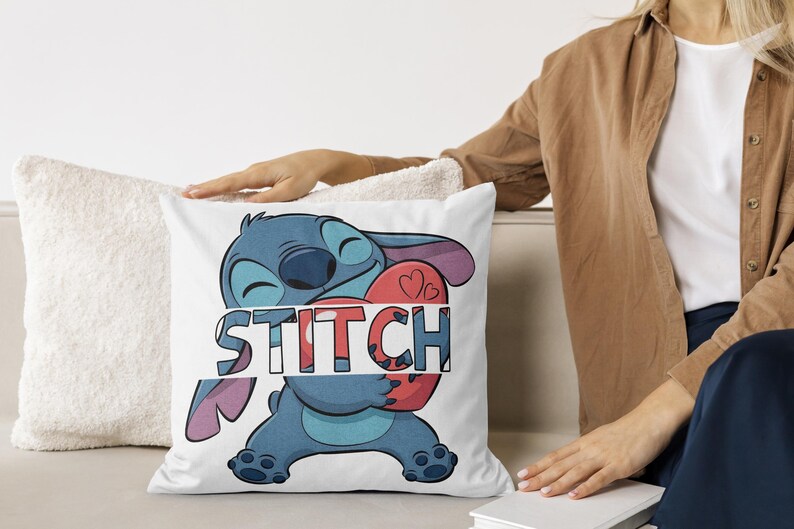 May include: White square pillow featuring a blue cartoon character holding a red heart with two small hearts. The word "STITCH" is printed across the character in a colorful, blocky font. The pillow is on a white couch.